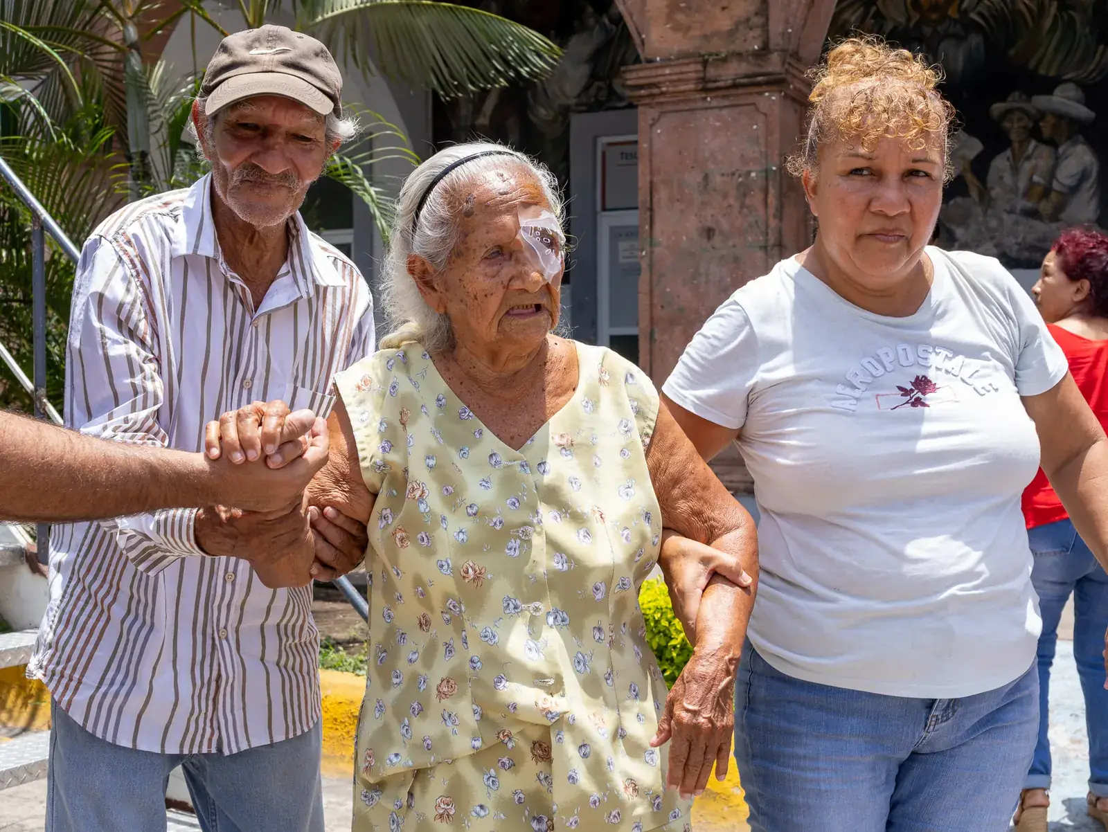 Paciente mayor con protector en el ojo camina sostenida por un hombre y una mujer